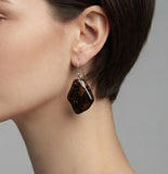 A close-up of a model's ear wearing a dangling earring with an irregular polished dark brown amber and a silver fishhook backing, set against a plain gray background