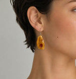 A close-up of a model's ear wearing a dangling amber drop earring , against a plain light gray background.