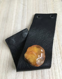 A black leather cuff bracelet featuring a large, organic-shaped amber stone set on the front, displayed on a light wood background.