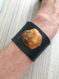 A wide black leather cuff bracelet worn on a wrist, featuring a large, organic-shaped polished amber stone .