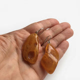 Two polished amber stone drop earrings with silver fishhook wires, held in a person's hand against a plain white background.