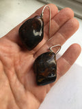 A pair of handmade tumbled  amber stone drop earrings with silver fishhook wires, held in a person's hand. The stones are dark brown and black with natural vegetable inclusion.